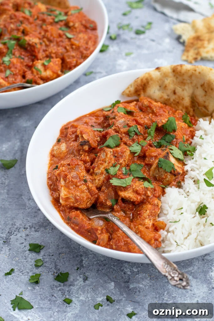 A fork in a large white bowl of chicken tikka masala next to a large scoop of rice and sprinkled with cilantro.