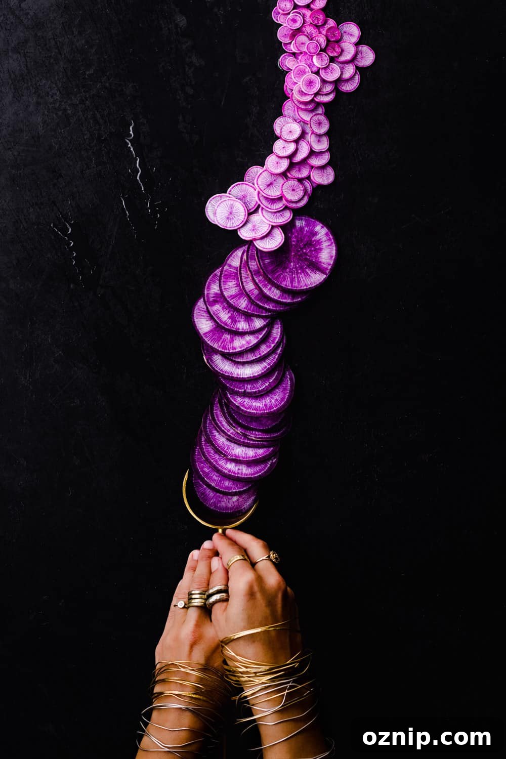 Hand holding a gold spoon filled with sliced purple radish varieties arranged in a gradient, overhead view on a dark background.