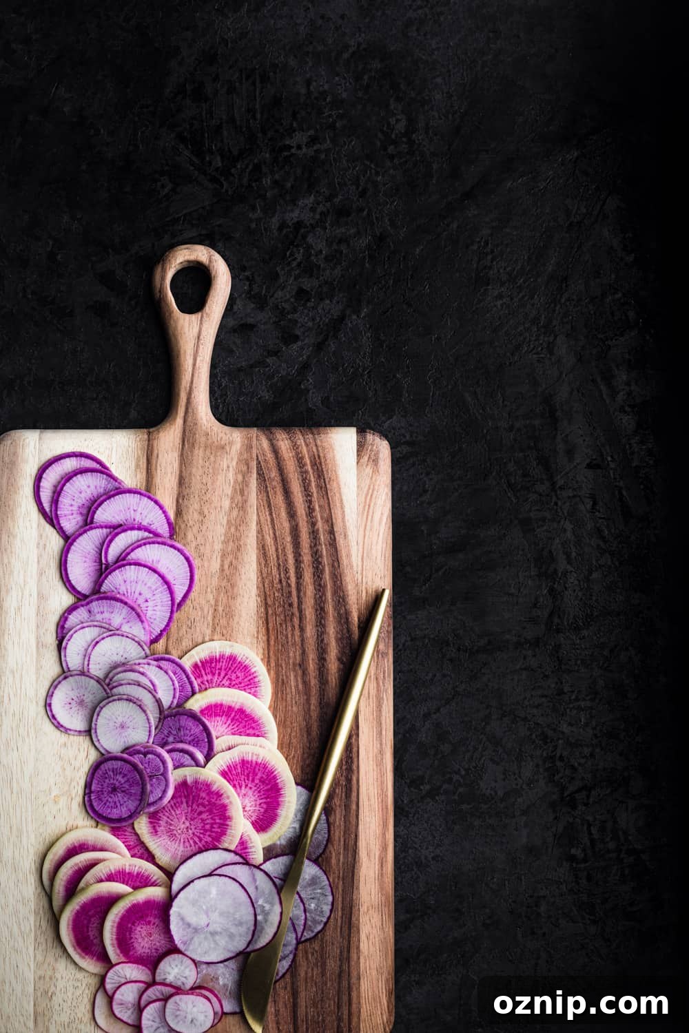 Thinly sliced pink and purple radish varieties on a wooden cutting board with a gold knife, overhead on a dark background.