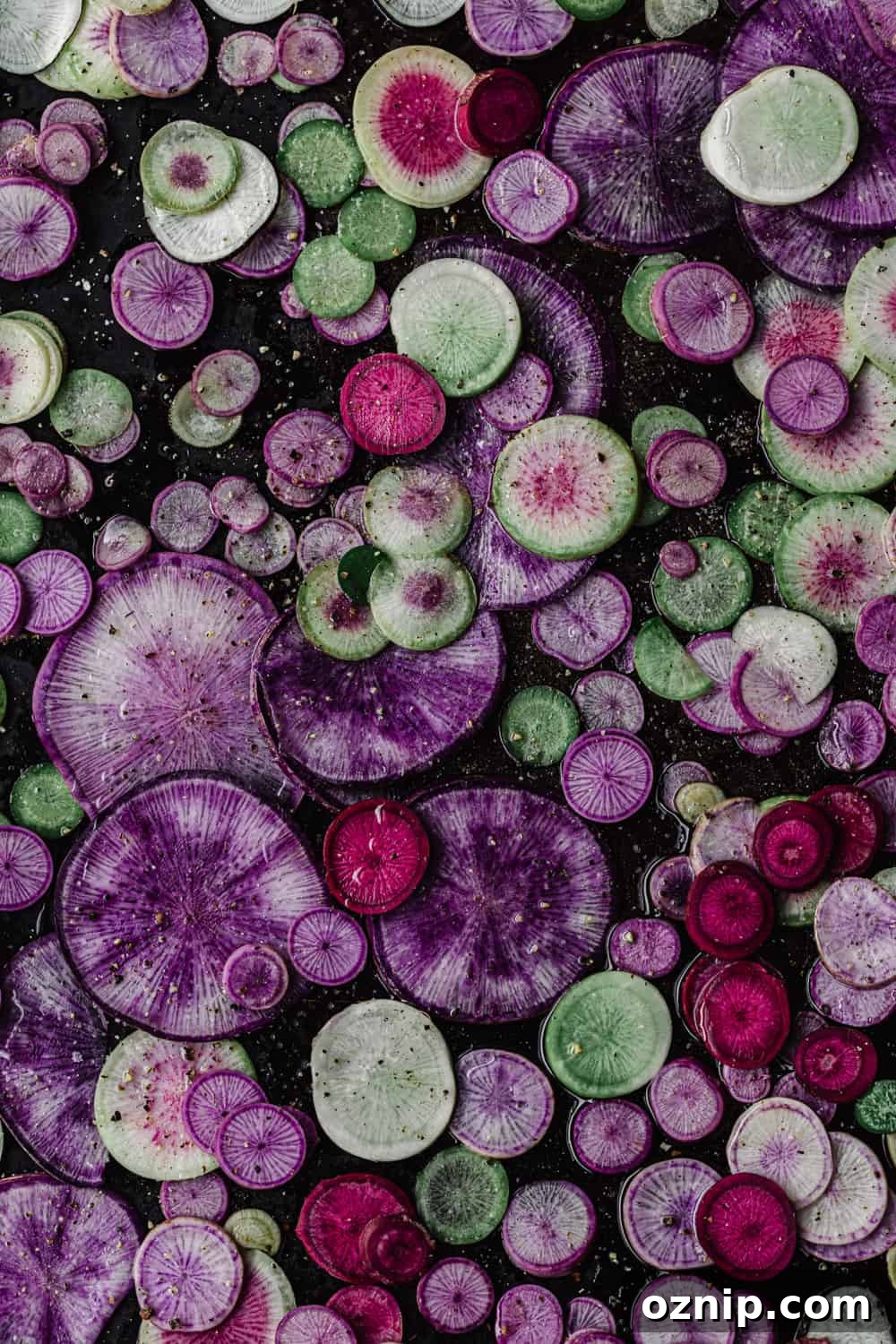Thinly sliced pink, purple and green radishes on a baking sheet before roasting, overhead view.