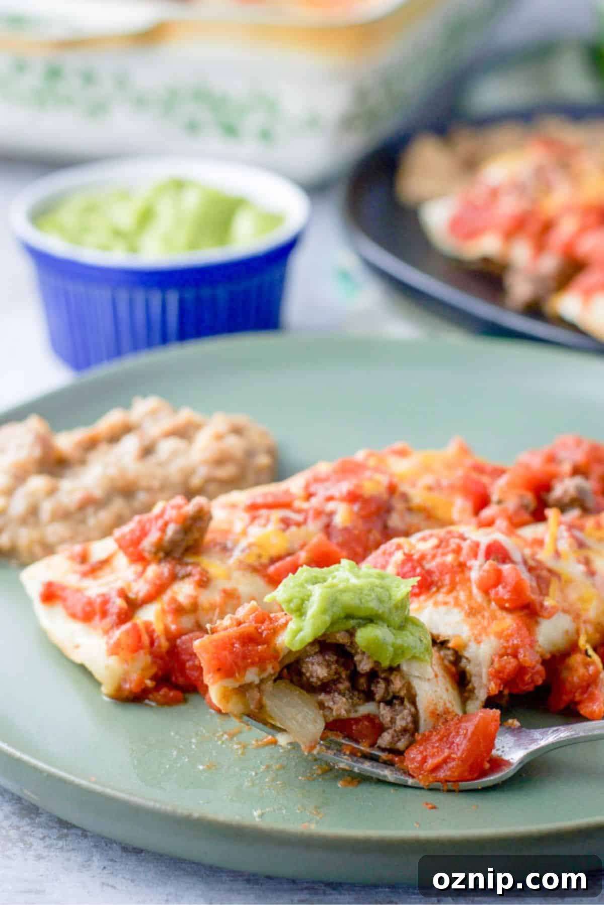 Cheesy Ground Beef Enchiladas with Red Sauce 2 A fork with a bite of enchilada resting on a plate with two enchiladas and beans. There is guacamole and more enchiladas in the background