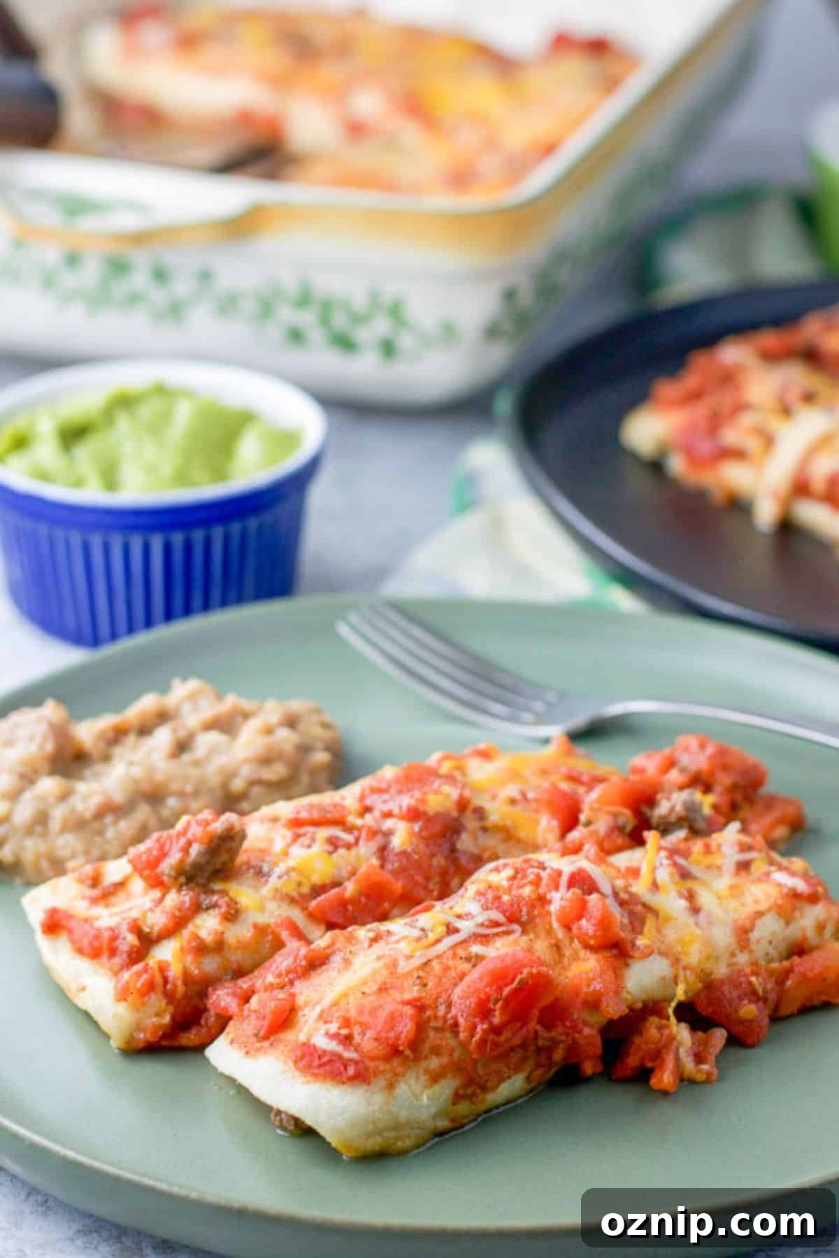 Cheesy Ground Beef Enchiladas with Red Sauce 8 Two enchiladas with a red sauce on a plate with some beans. There is some guacamole in the background along with another plate of enchiladas