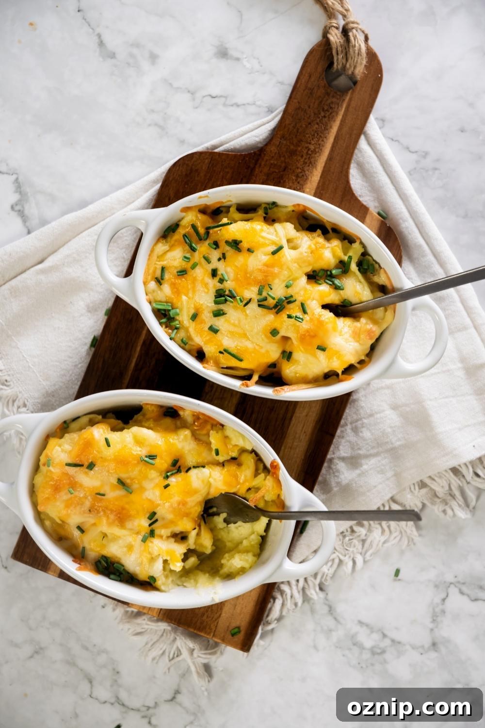 Two white casserole dishes on top of a wooden cutting board with cheesy mashed potatoes in it.