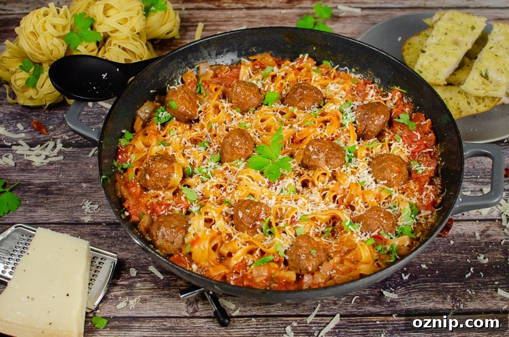One-Pot Spicy Meatball Pasta served in a cast iron pan with sprinkled chopped parsley on top for garnish, with garlic bread served on the side on a grey Parmesan cheese with a silver grater, black spoon to serve the food and dry balls of tagliatelle in the background of the picture