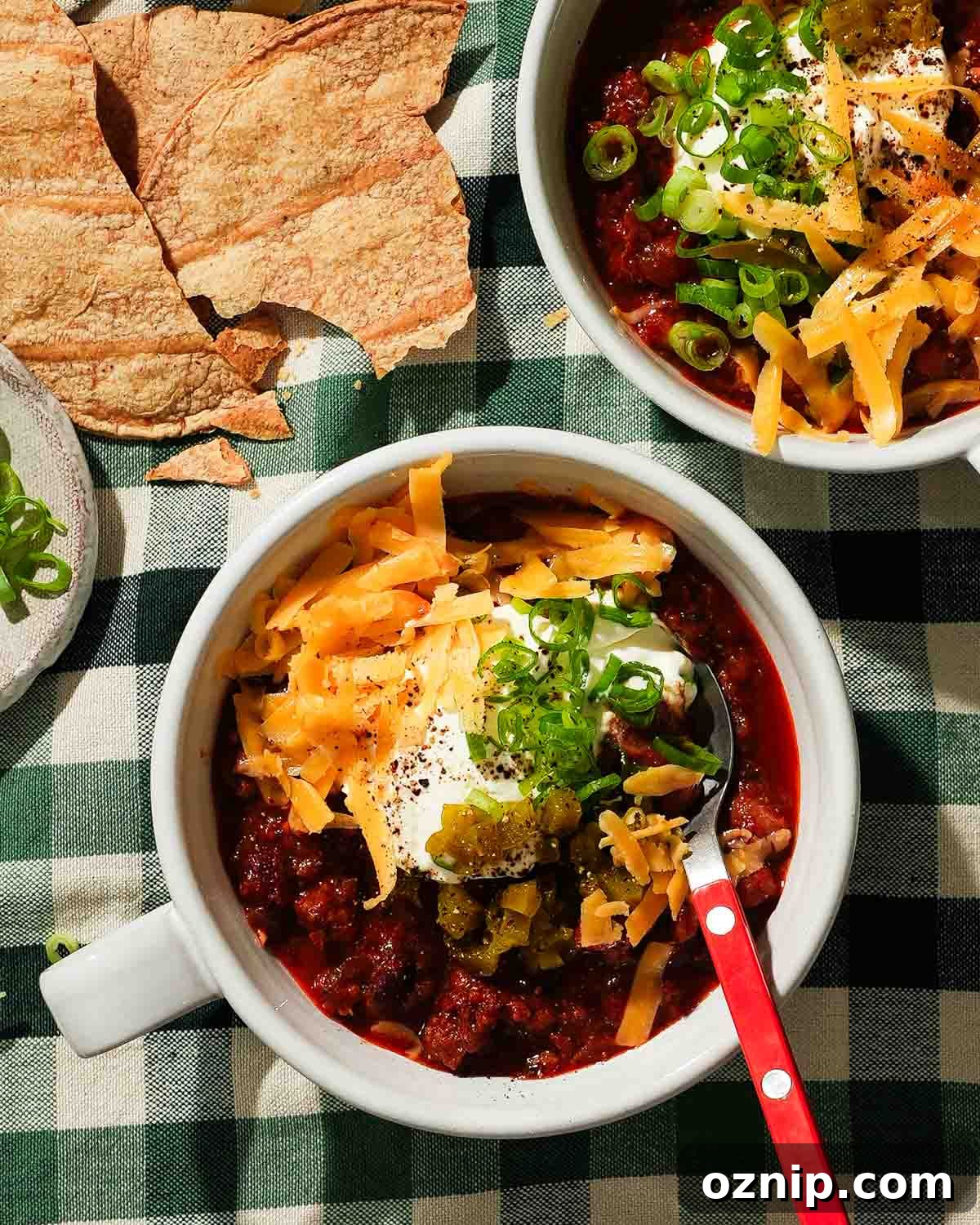A bowl of chili topped with shredded cheese, pickled jalapeños, and green onions with an orange spoon.