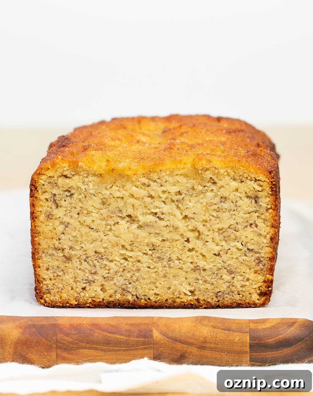 partially sliced loaf of light brown banana bread with brown flecks on white paper on wooden cutting board