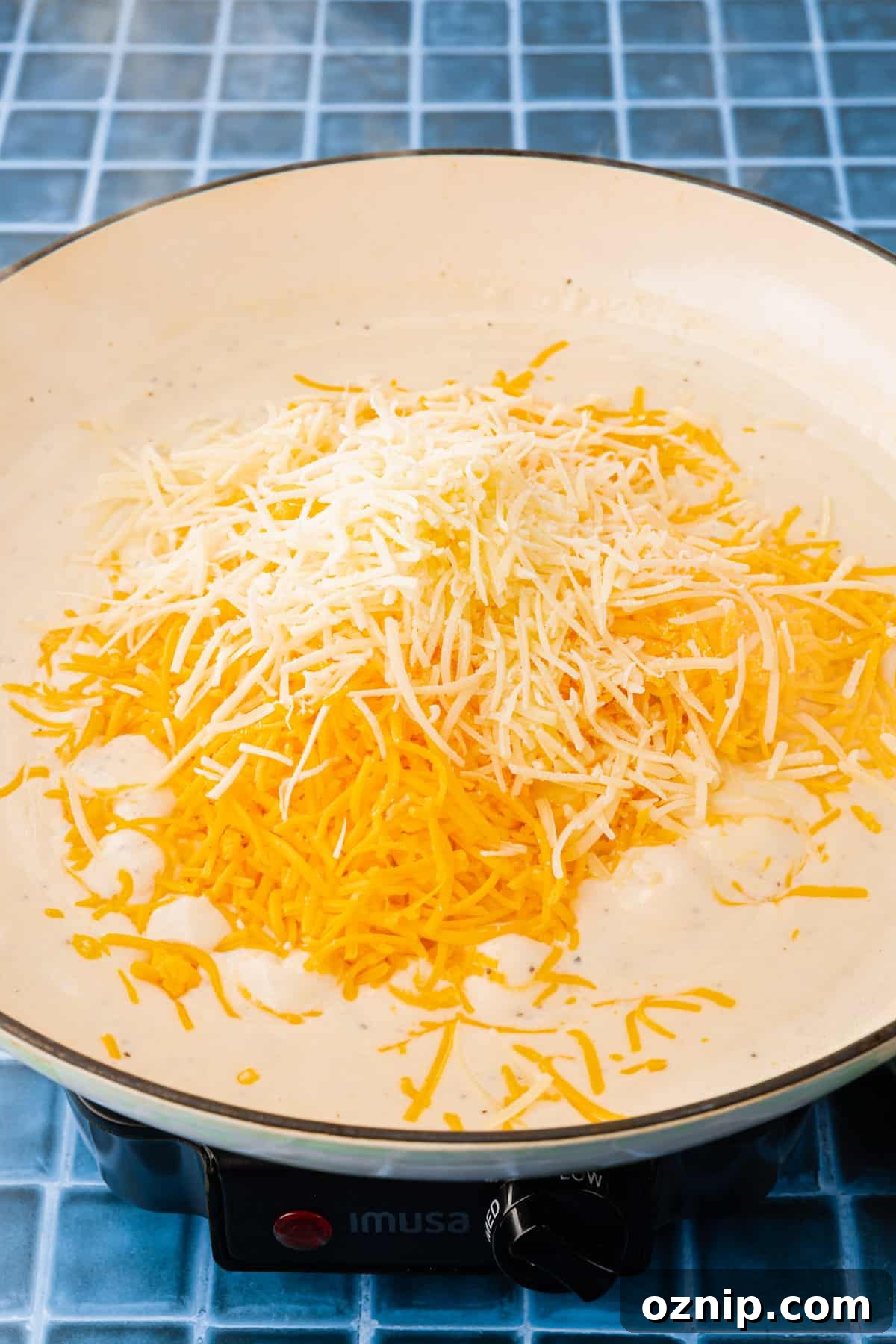 Creamy Gluten Free Mac and Cheese Recipe 8 A white braising pan with a white creamy sauce topped with shredded cheddar cheese and parmesan cheese before mixing together.