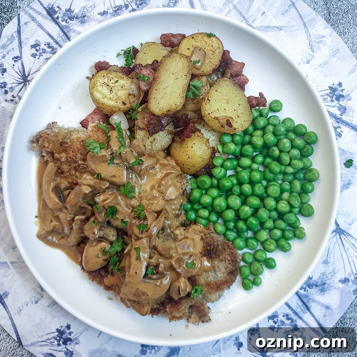 Overhead shot of a crispy beef schnitzel on a white plate with peas, potatoes and mushroom sauce.