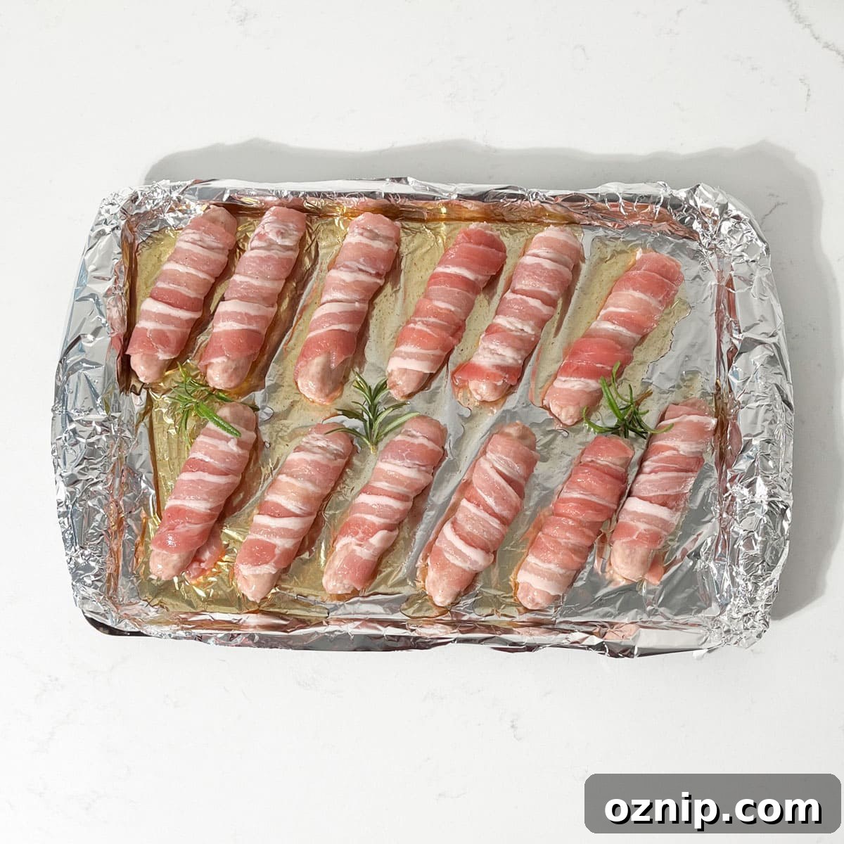 Whiskey Honey Glazed Pigs in Blankets 14 Pigs in blankets on a baking tray, covered in glaze and with pieces of rosemary between the sausages.