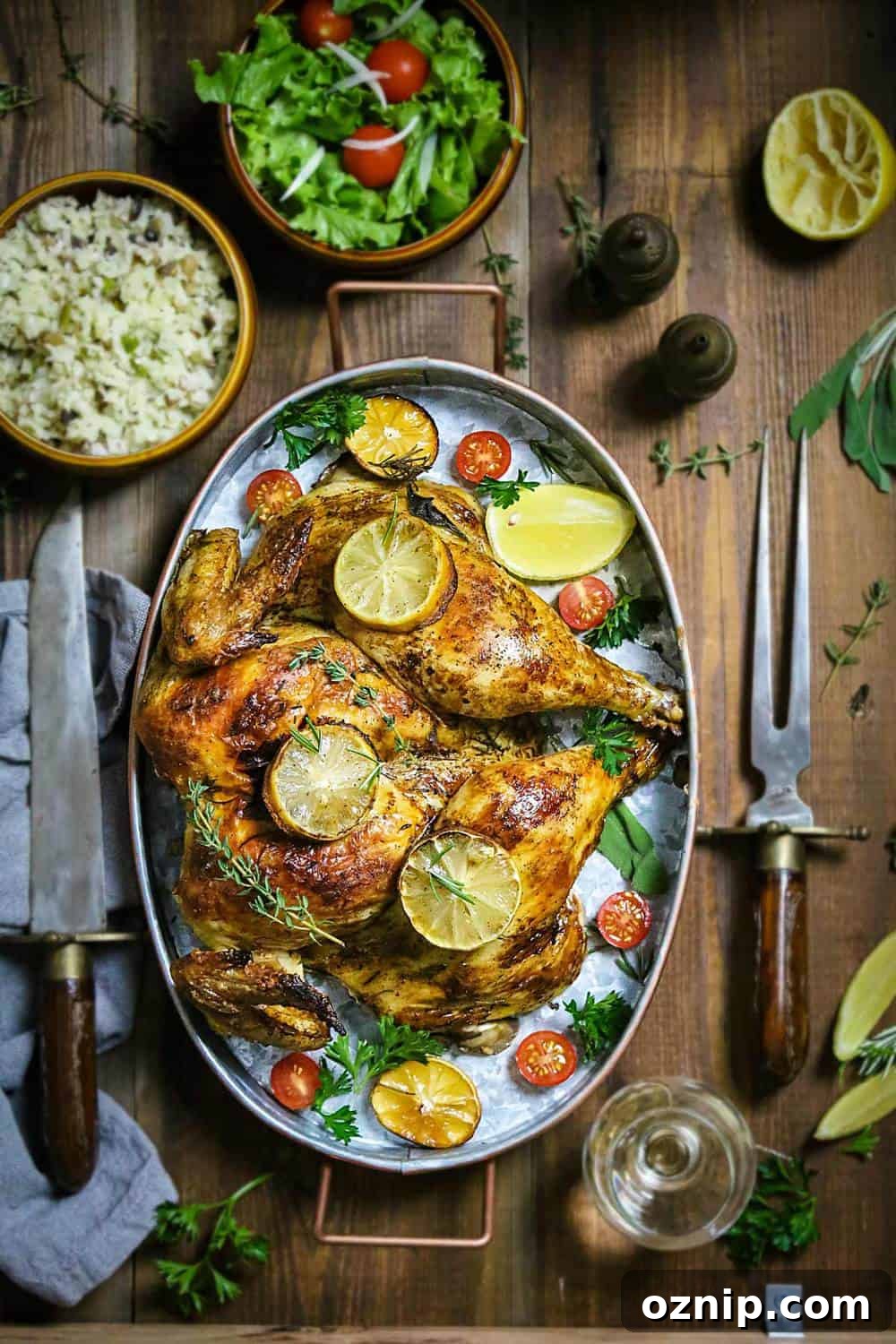 An oval dish with roasted whole chicken with lemon slices and fresh herbs sitting on a wooden table with cutlery and side dishes.