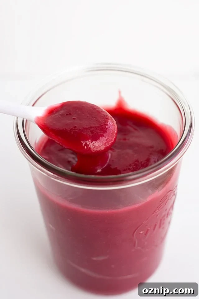 Mixed Berry Curd Recipe 2 A jar filled with smooth, dark red homemade fruit curd, with a white spoon holding a spoonful above the jar.