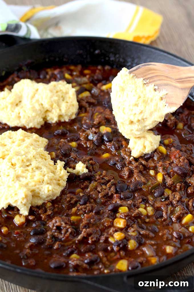 Raw Cornbread is dolloped over a mixture of ground beef chili with beans and corn, using a wooden spoon.