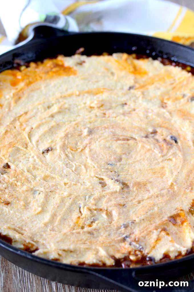 View of a skillet sitting on a wooden surface containing raw cornbread mixture spread over the top of chili. 