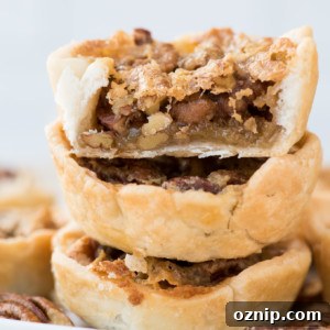 A close-up of three stacked mini pecan pies, with the top tart bitten to reveal the gooey, nut-filled filling inside the flaky pastry crust.