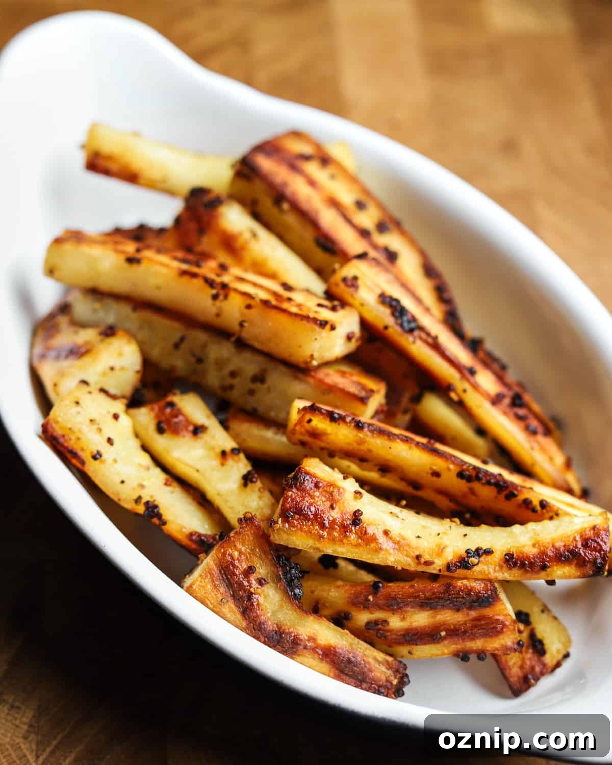 Photo of roast parsnips in a white oval oven dish placed on a wooden table.