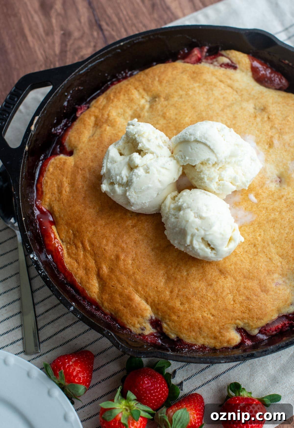A cast iron skillet is sitting on a table filled with strawberry sourdough cobbler and three scoops of ice cream on top.