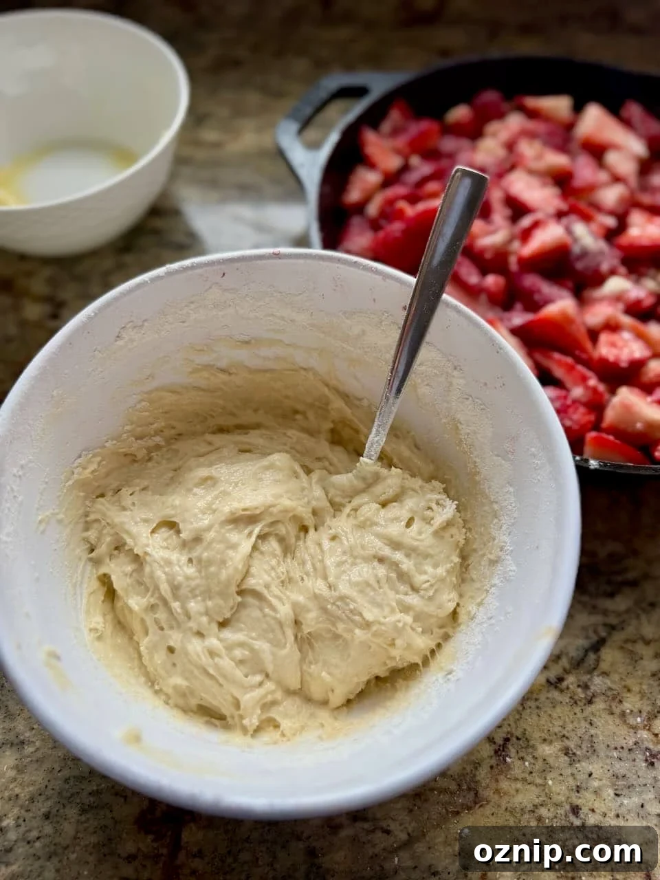A bowl of cobbler topping batter sits next to a skillet filled with strawberries.