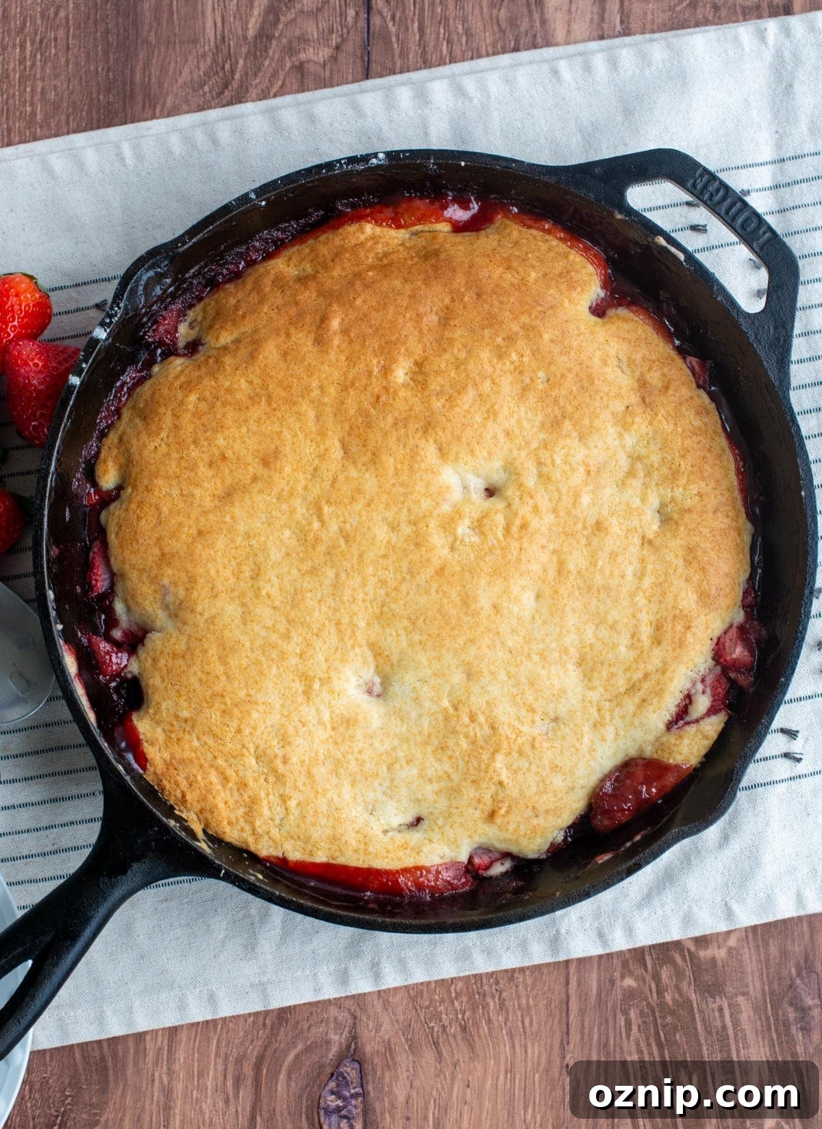 A golden brown sourdough discard cobbler in a cast iron skillet sits on a tea towel next to some fresh strawberries.