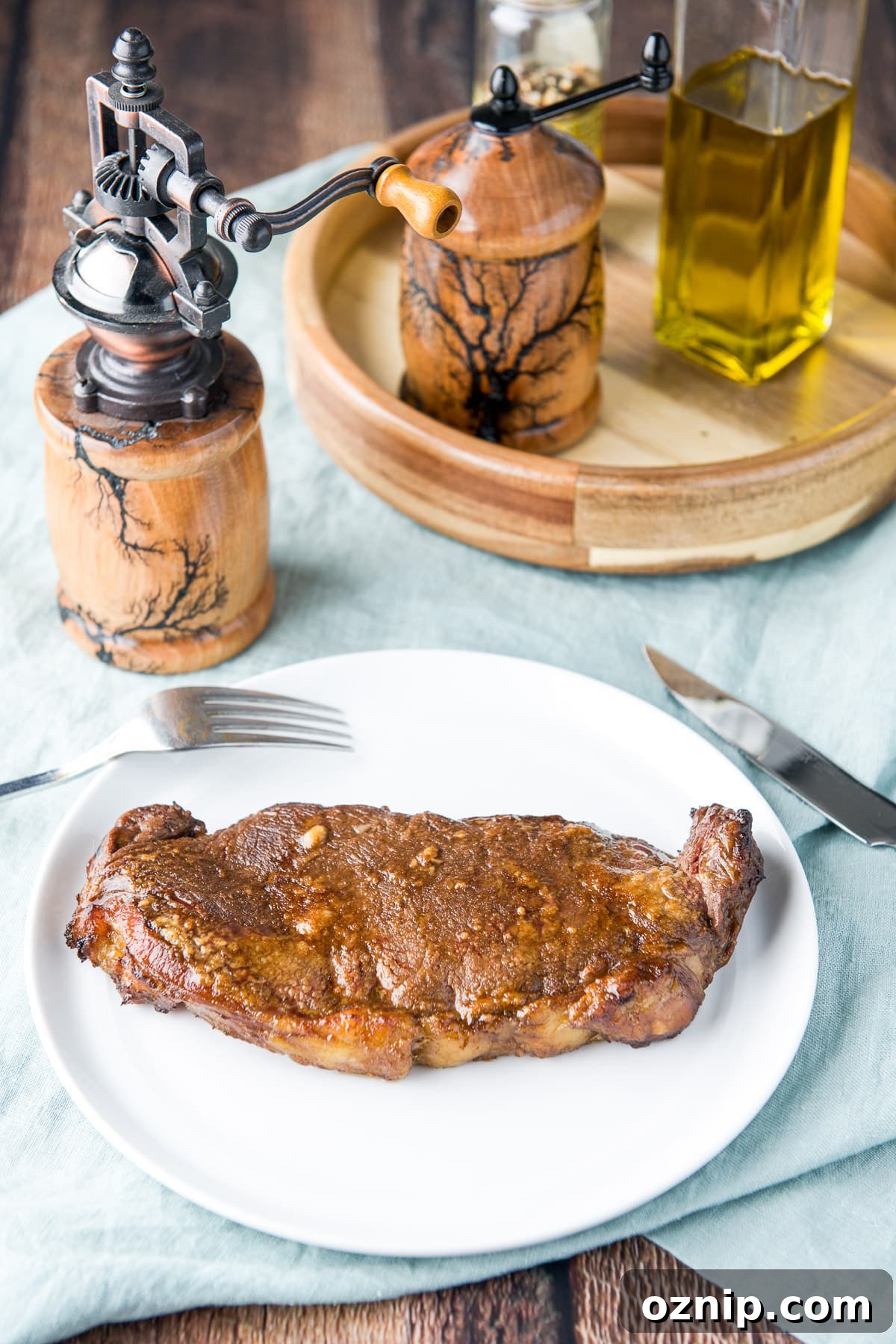 A white plate with steak on it along with a fork and knife with salt and pepper mill on the table