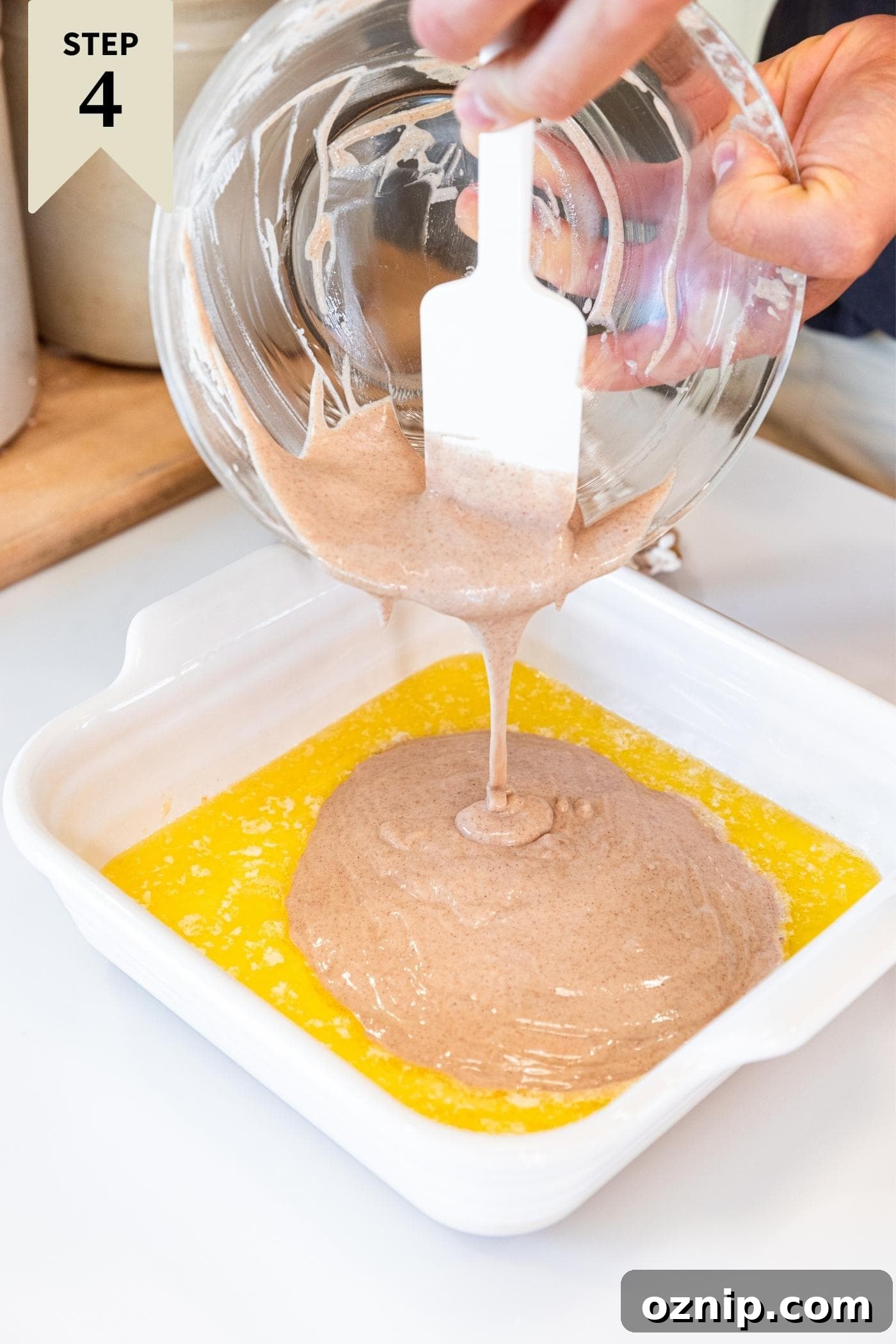 Hand scooping liquid batter into white baking dish filled with melted butter all on white countertop.