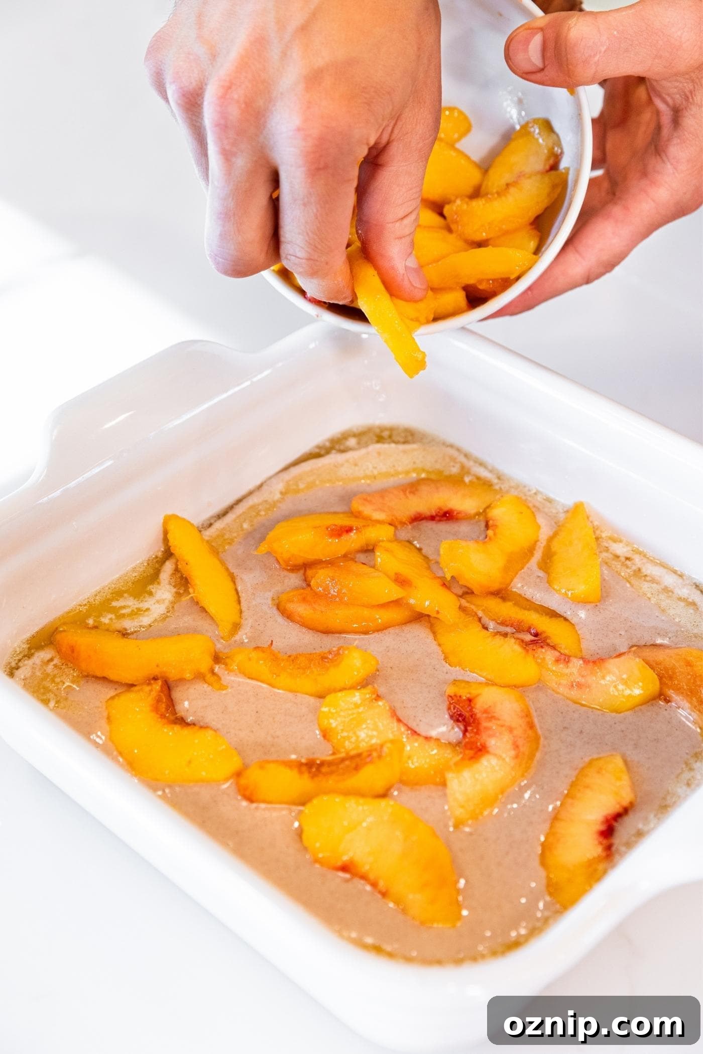 Hand sprinkling peach slices on top of light brown batter in white baking dish on white countertop.