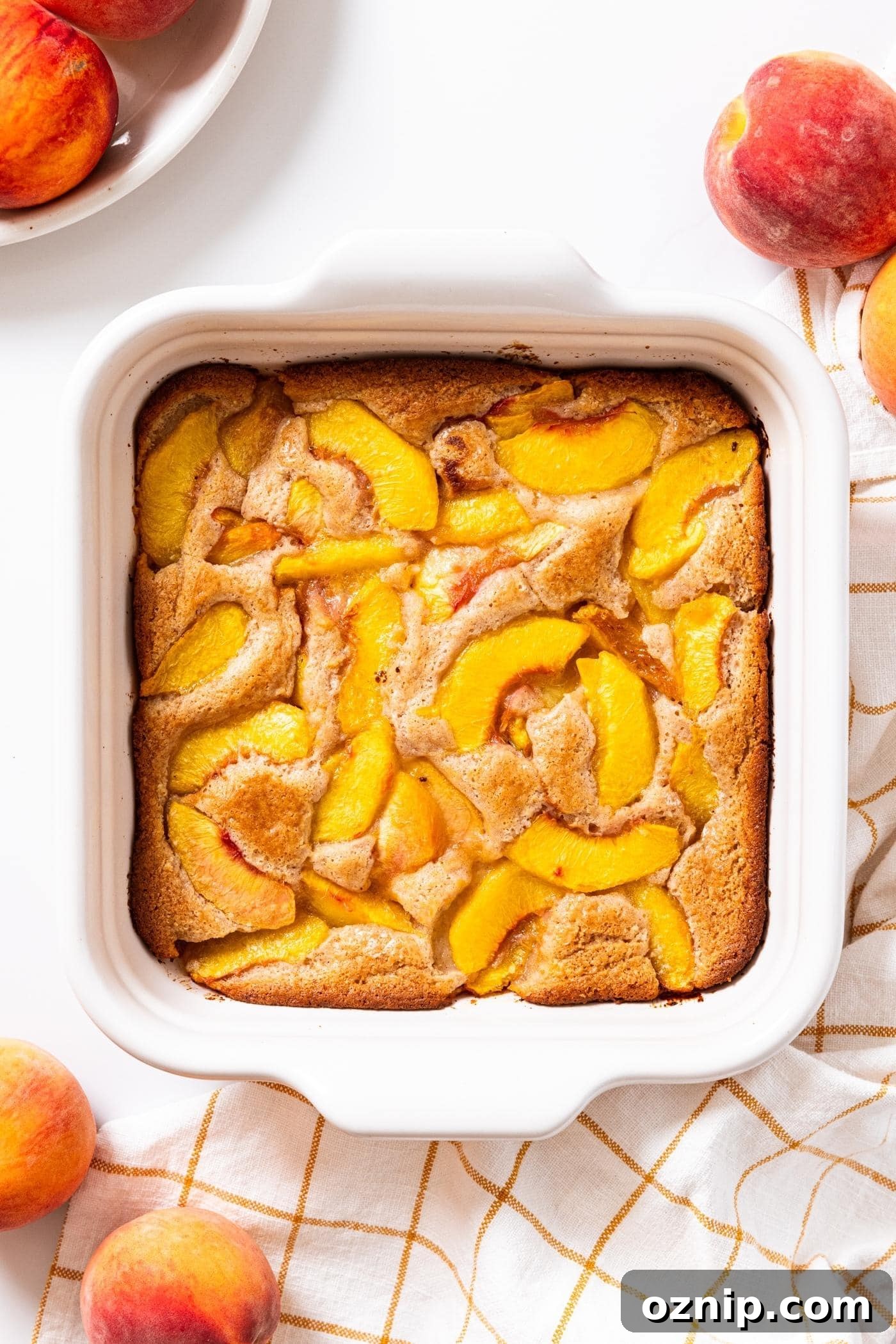 White baking dish sitting on white countertop filled with baked lazy peach cobbler surrounded by unsliced whole peaches.