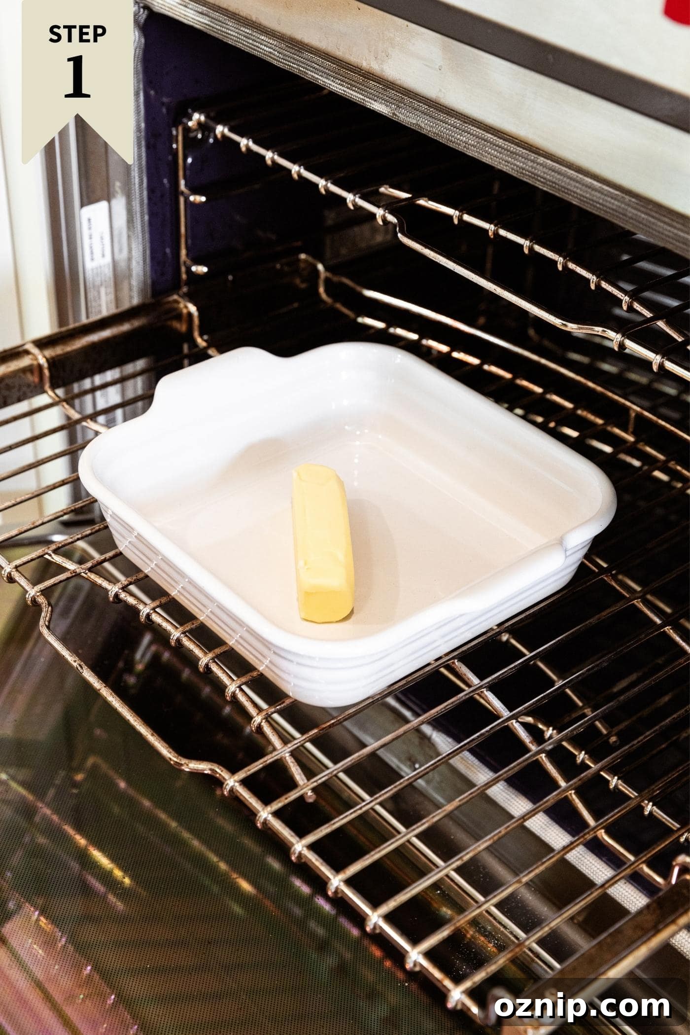 White baking dish filled with unmelted stick of butter sitting on metal rack in preheating oven.