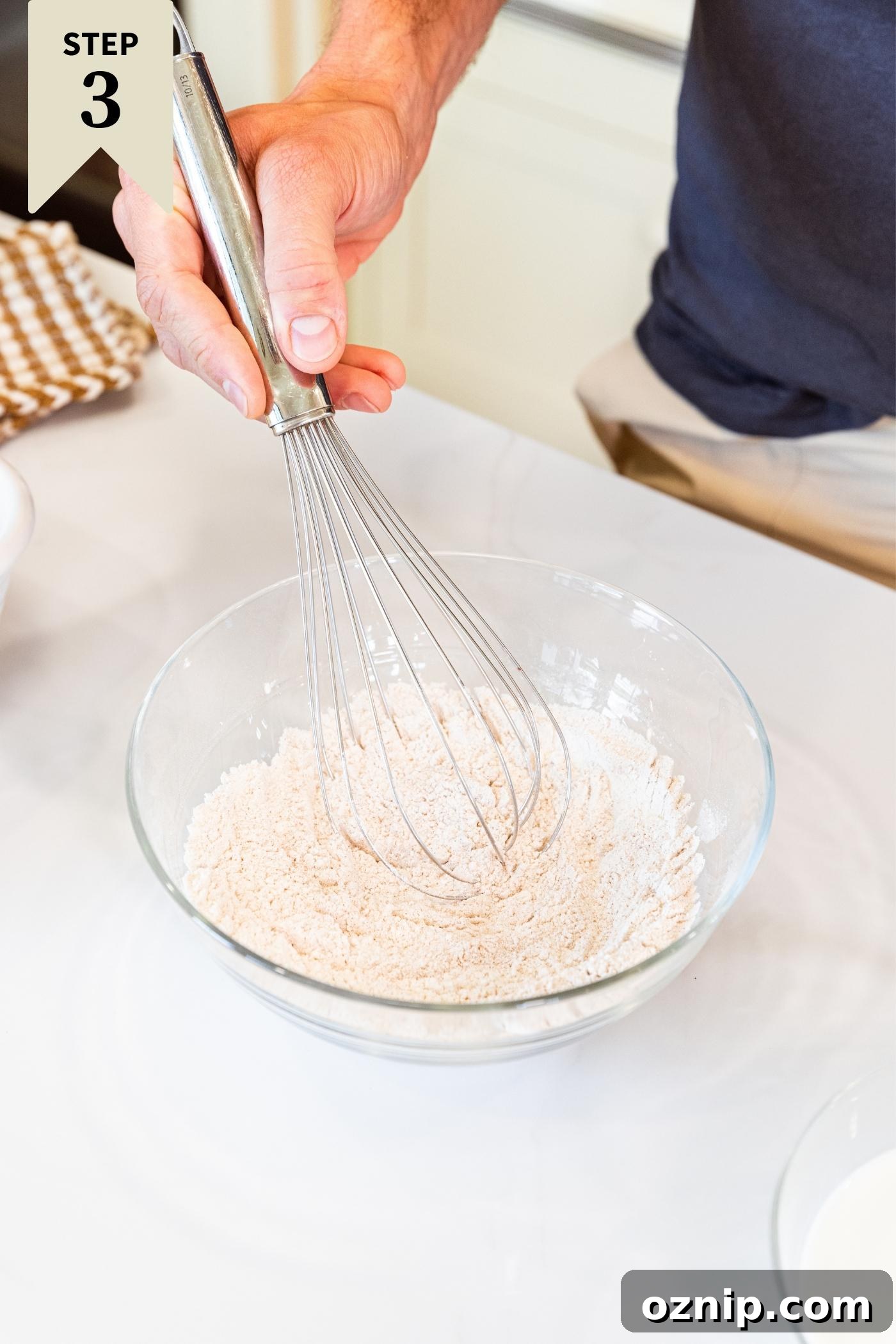 Hand holding whisk mixing flour and other dry ingredients in glass bowl on white countertop.