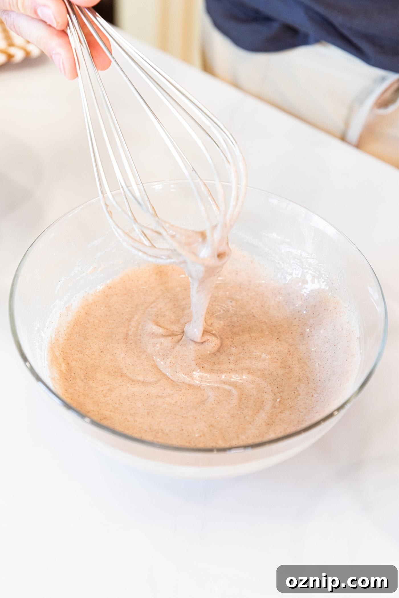 Hand holding whisk with liquid batter that's light brown in color on white countertop.