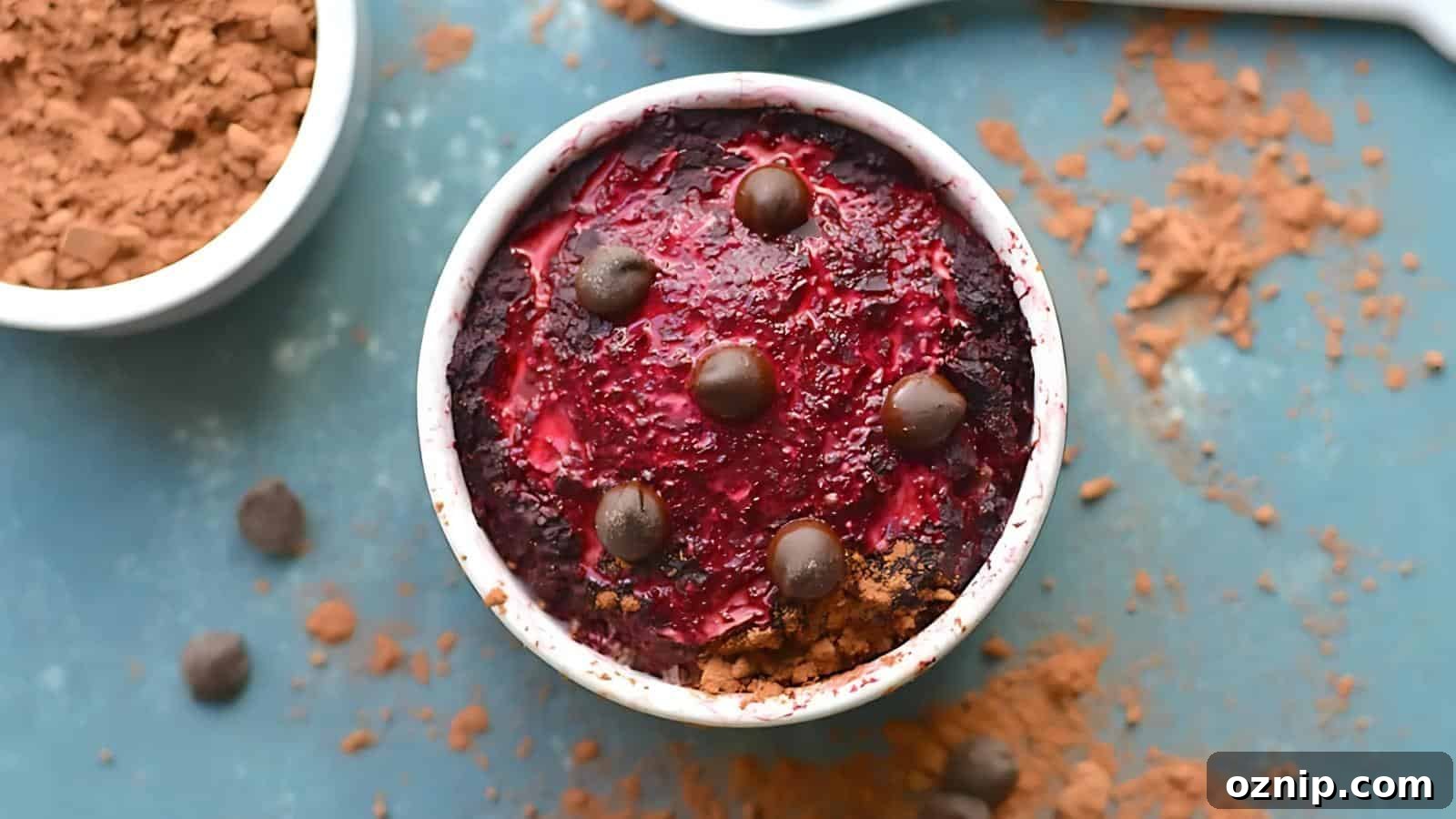 A small white ramekin filled with a baked dessert topped with berry sauce and chocolate pieces, surrounded by cocoa powder and chocolate chips on a blue surface.
