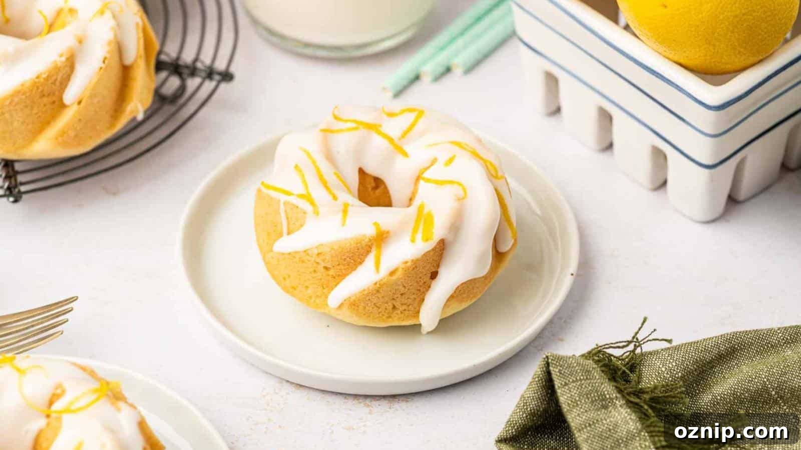 A small Bundt cake with white icing and lemon zest sits on a white plate, with lemons, a glass of milk, and candles in the background.