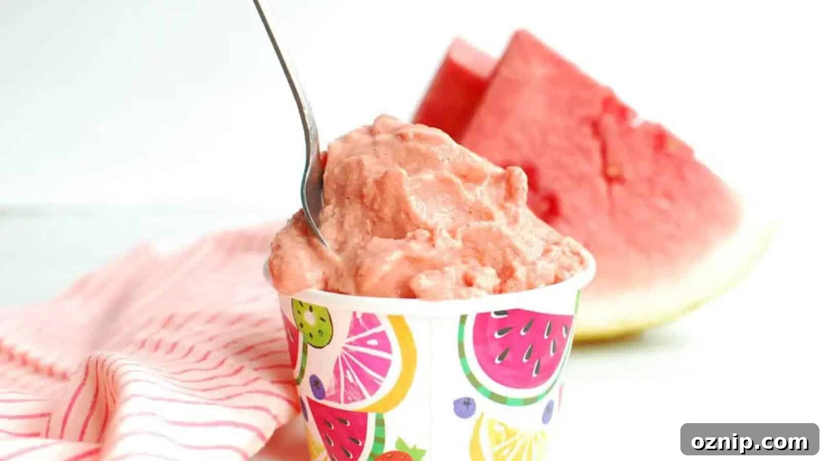 A cup of pink watermelon sorbet with a spoon, set on a striped cloth, with a slice of watermelon in the background.