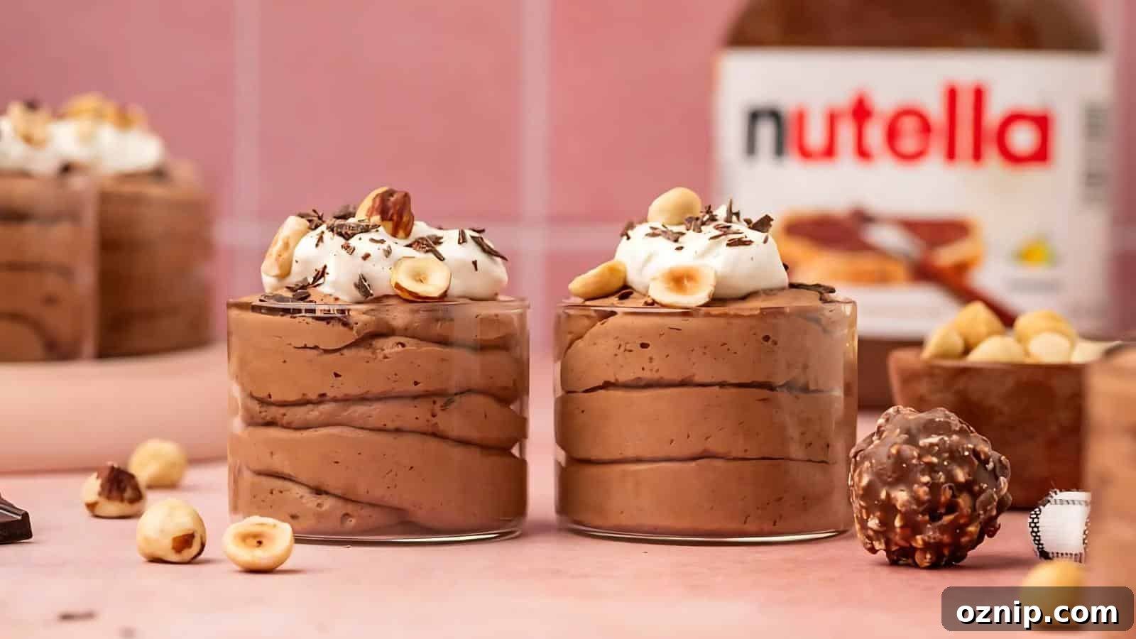 Two glass cups filled with chocolate mousse topped with whipped cream, hazelnuts, and chocolate shavings, with a Nutella jar and hazelnut chocolates in the background.