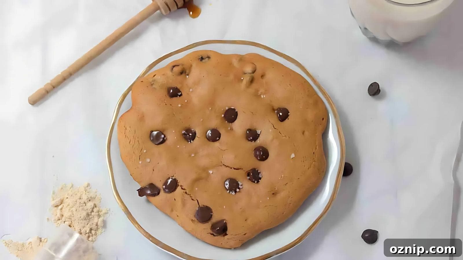 A large chocolate chip cookie on a white plate, surrounded by chocolate chips, flour, a honey dipper with honey, and a glass of milk on a white surface.
