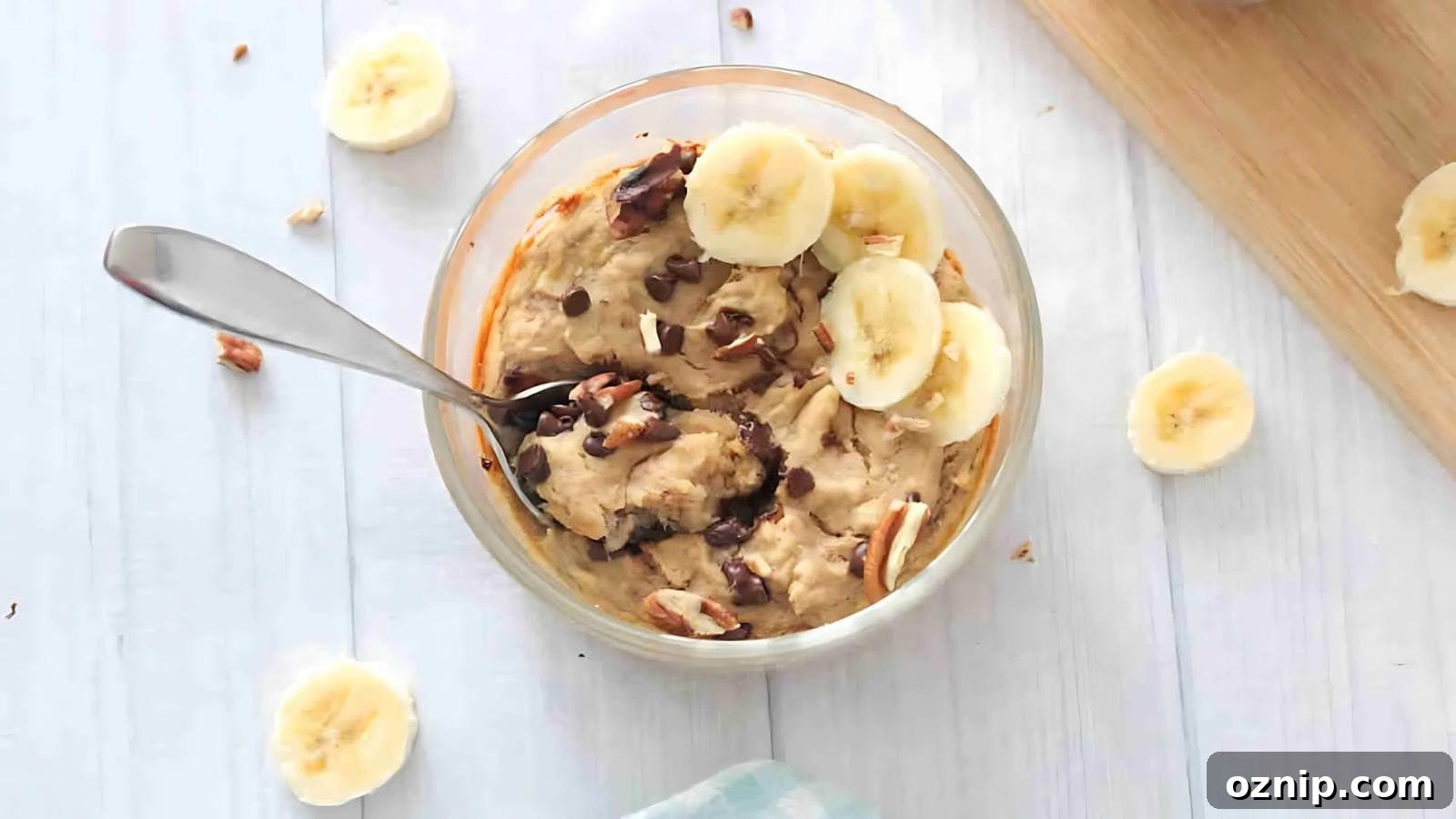A glass bowl of oatmeal topped with banana slices, nuts, and chocolate chips, with a spoon inside, on a light wooden surface.