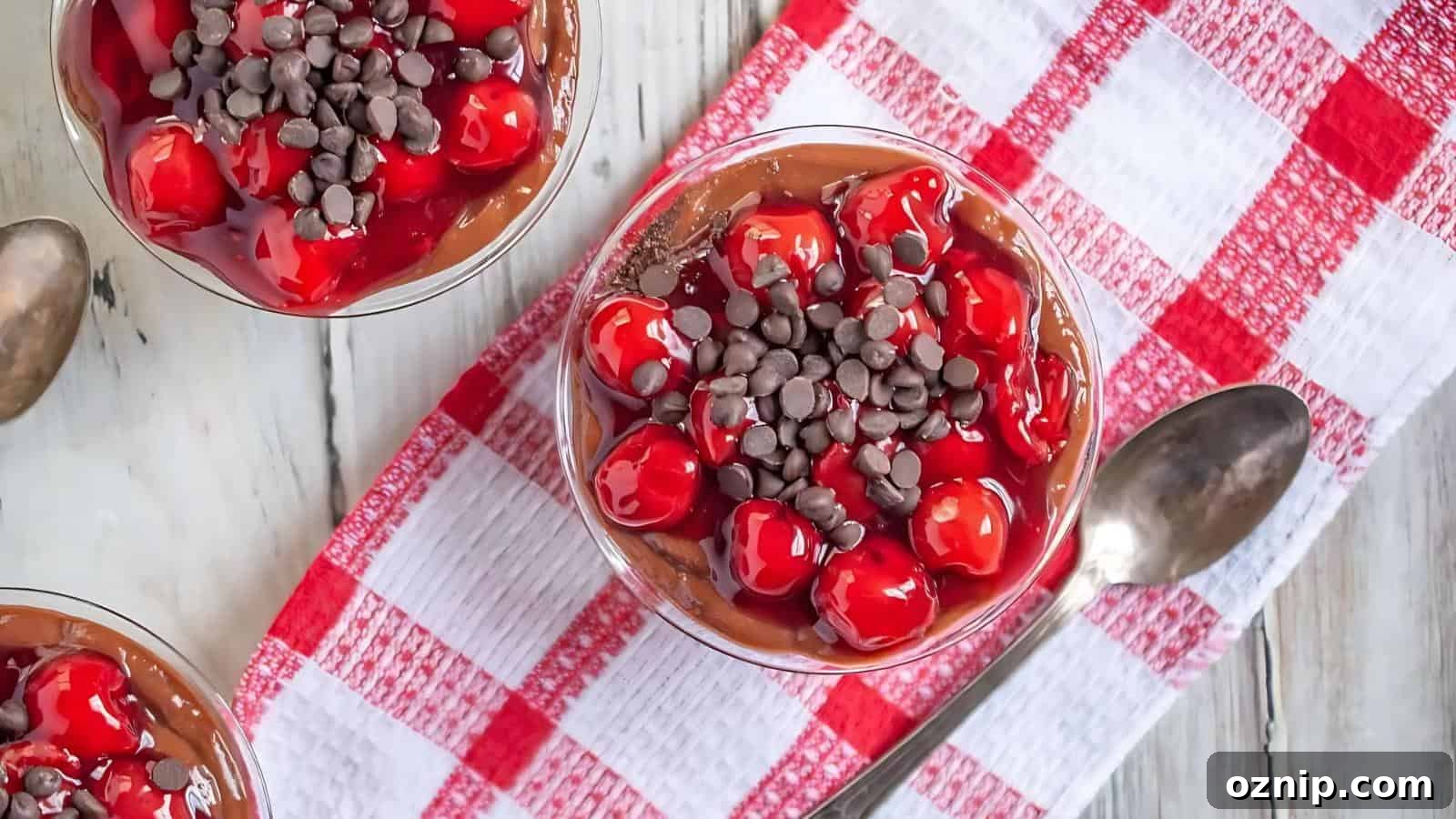 A glass bowl of chocolate pudding topped with cherries and chocolate chips, next to a spoon on a red and white checkered cloth.