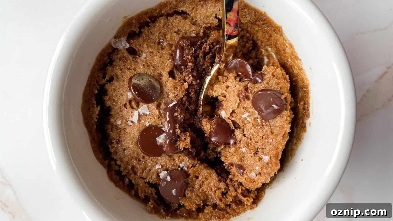 A chocolate chip cookie mug cake with melted chocolate chips and a sprinkling of sea salt, partially scooped with a spoon in a white ramekin.