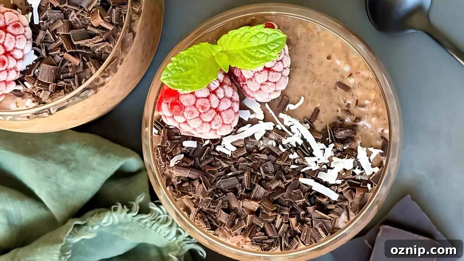 A glass bowl of chocolate chia pudding topped with frozen raspberries, mint leaves, shredded coconut, and chocolate shavings, with a spoon and green cloth nearby.