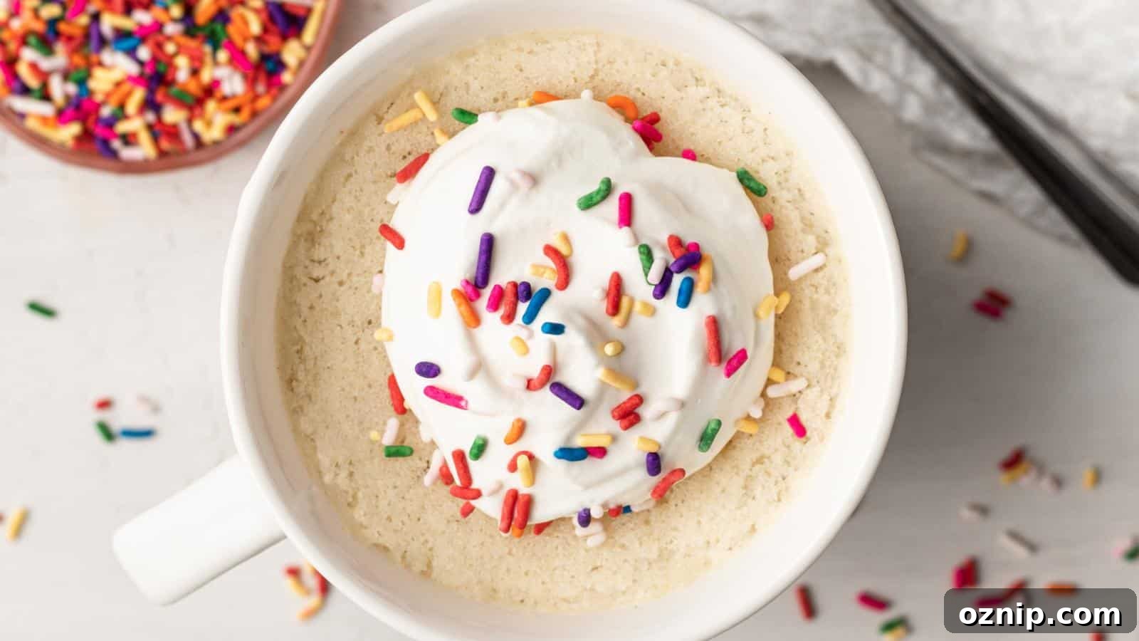 A mug cake topped with whipped cream and colorful sprinkles sits on a white surface, with extra sprinkles scattered nearby.