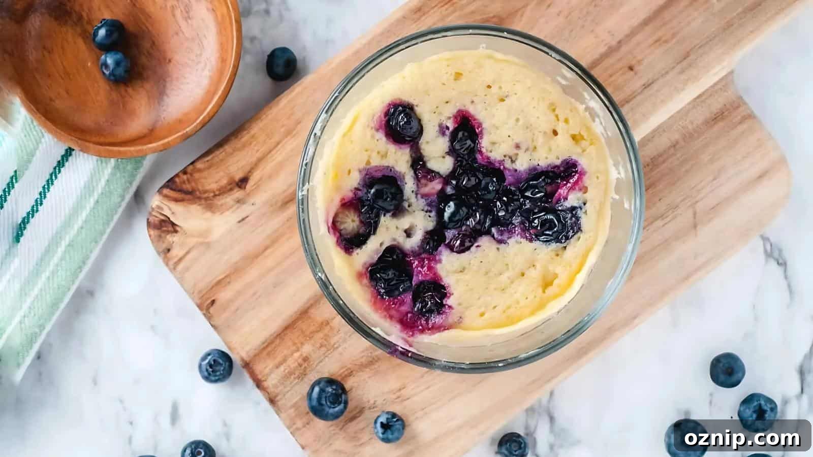 A glass bowl with a blueberry mug cake sits on a wooden board, surrounded by fresh blueberries and a wooden dish on a marble surface.