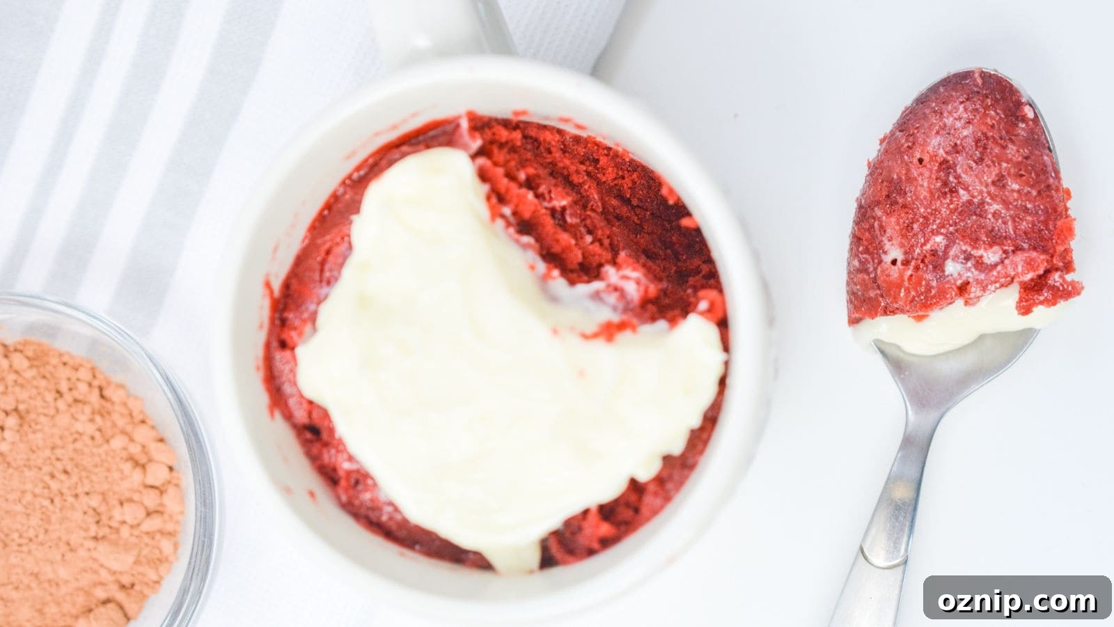 Red velvet cake with a layer of cream cheese frosting in a white ramekin, next to a spoon with a bite taken out, on a white background.