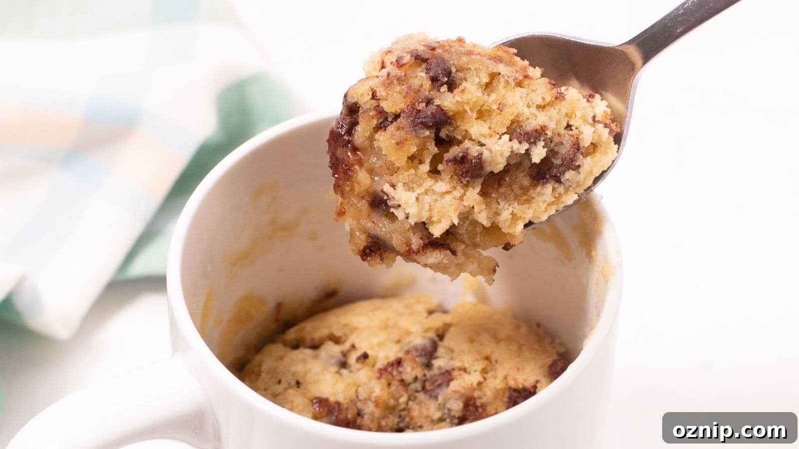 A spoon lifting a bite from a chocolate chip mug cake inside a white mug.
