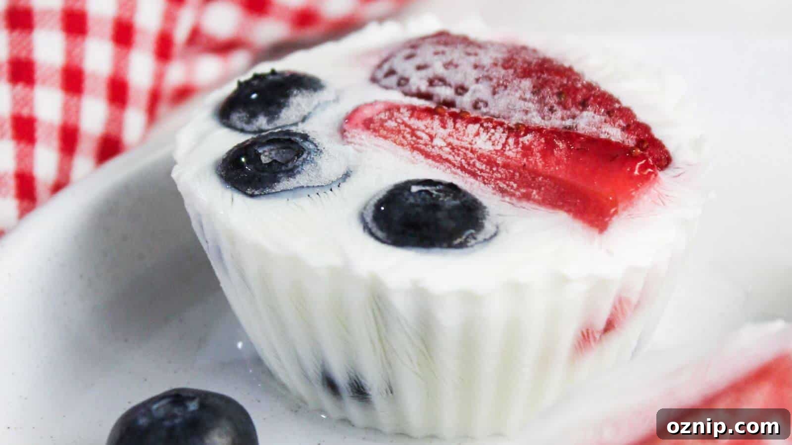 A close-up of a yogurt cup topped with blueberries and strawberry slices in a white silicone mold, placed on a white plate. A red and white checkered cloth is partially visible in the background.