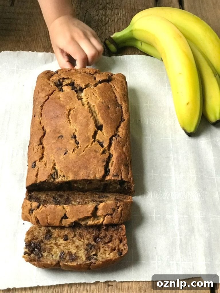 Overhead photo of a loaf of banana bread with a few slices already cut. There is a little hand sneaking in trying to take a chocolate chip from the bread. Three bananas are off to the side of the parchment lined wooden surface.