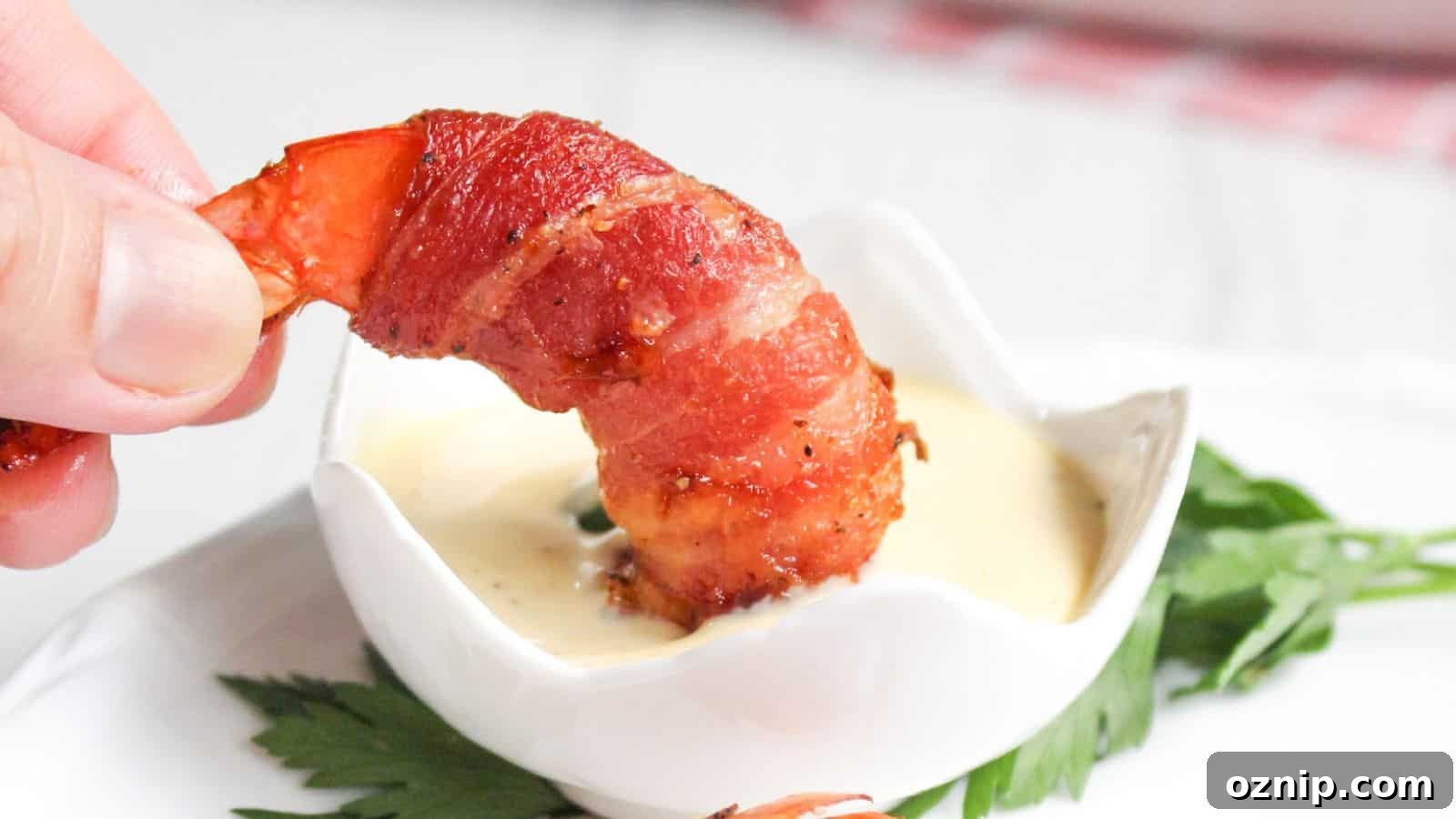 A hand holding a bacon-wrapped shrimp dipped in a small bowl of sauce, with a green leaf garnish on the side.