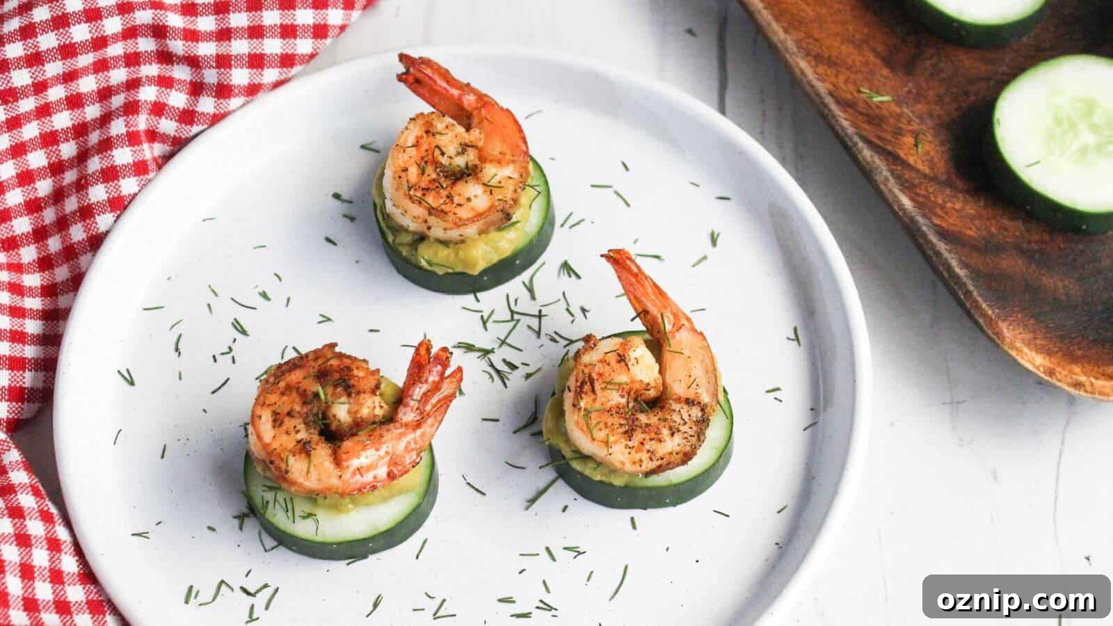 Three grilled shrimp atop cucumber slices garnished with herbs on a white plate with a red checkered napkin partially in view.