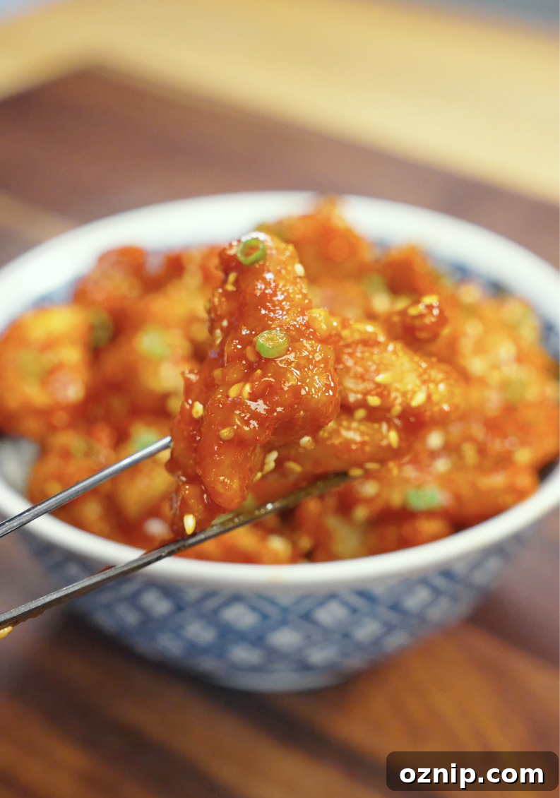 Close-up of a piece of Crispy Gochujang Chicken held with chopsticks, showcasing the thick sticky gochujang glaze, crunchy coating, and sesame seed garnish. Perfect for an easy Korean-inspired weeknight dinner.