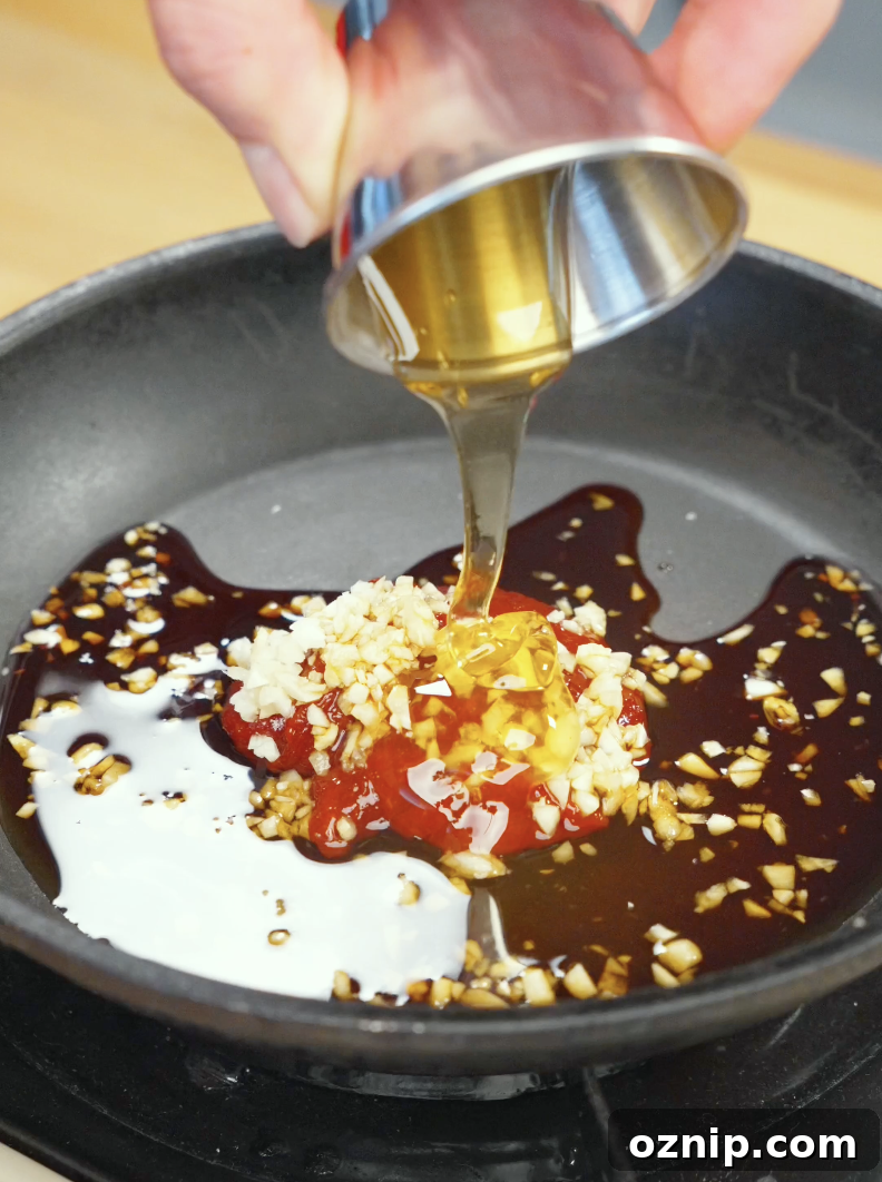 Honey being poured into a skillet with gochujang, garlic, soy sauce, rice vinegar, and sesame oil to create a sweet, spicy, and savory glaze for Crispy Gochujang Chicken Recipe.