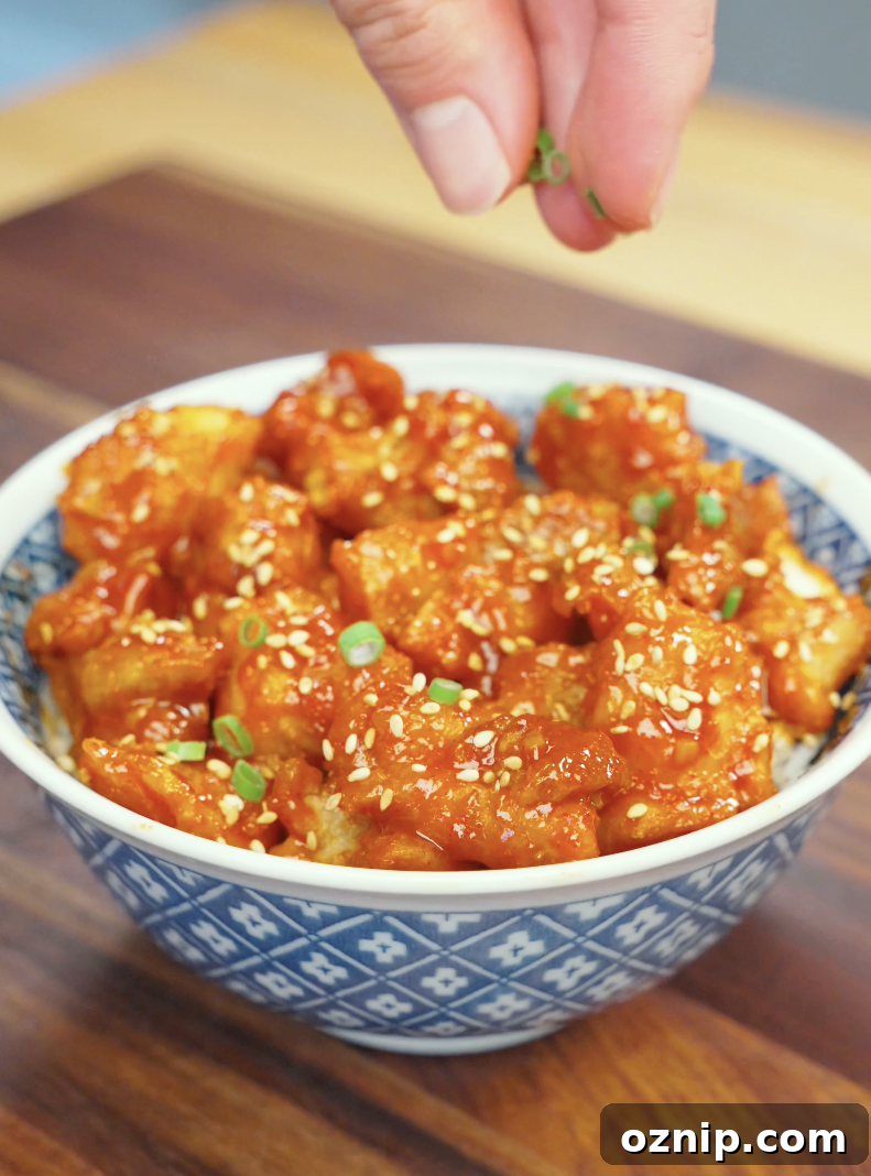 Fresh scallions being sprinkled over a bowl of Crispy Gochujang Chicken finished with sesame seeds and sticky gochujang glaze. This final garnish adds freshness, color, and texture to the dish.