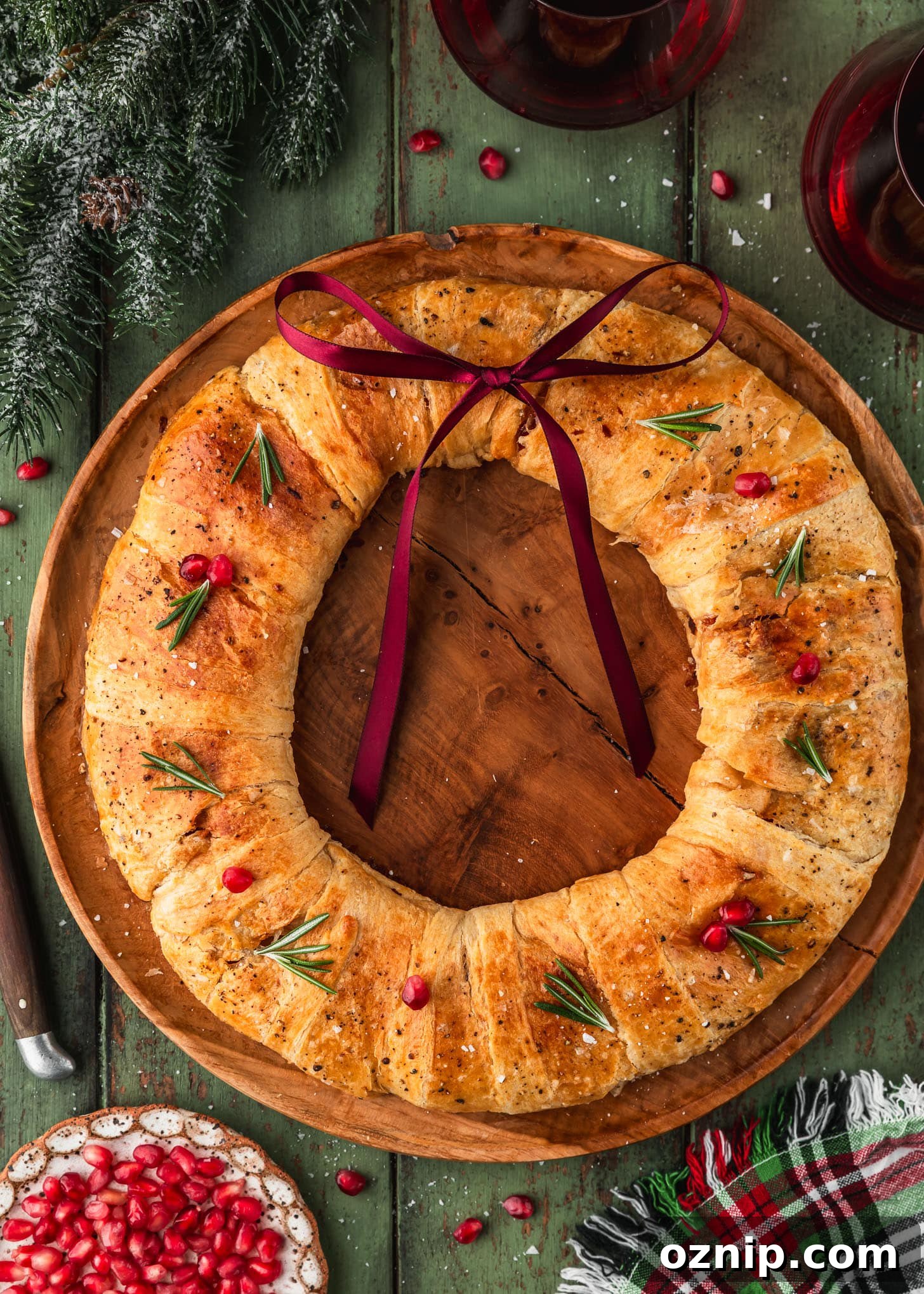 Flaky Bacon and Brie Crescent Wreath Recipe 2 A wood board with a bacon and brie crescent wreath on a green wood table next to garland, glasses of red wine, a plaid linen, and a white plate of pomegranate arils.